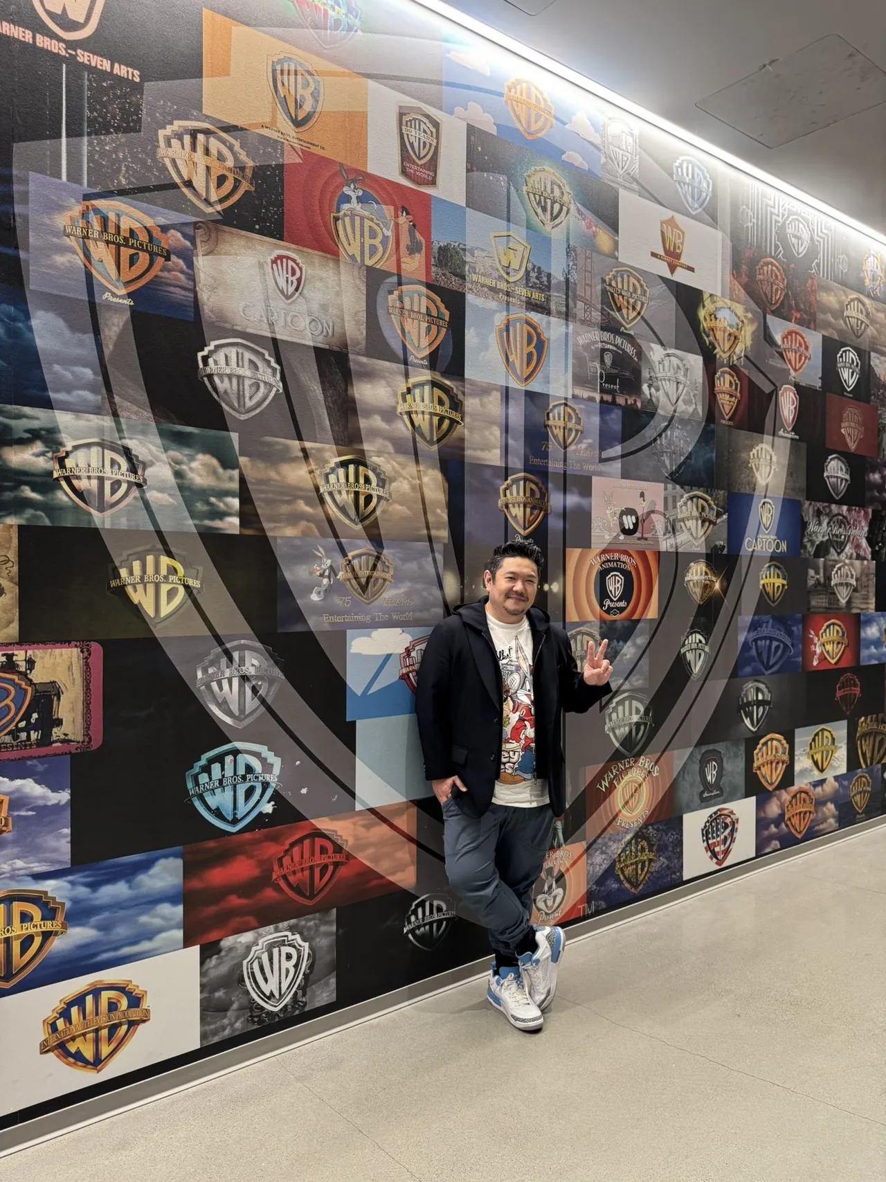 Hombre posando con el signo de la paz junto a un mural con logotipos de Warner Bros.